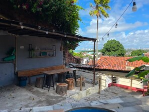 Un patio exterior con mesa y taburetes de madera, una piscina pequeña y vistas a un paisaje urbano.