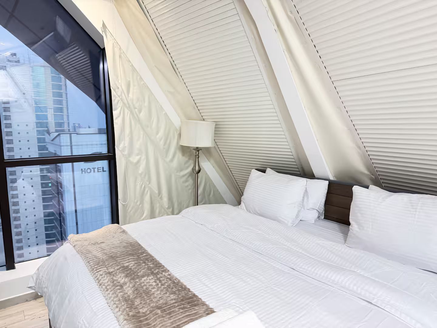 A hotel room with a large window, a bed with white bedding, and a lamp.