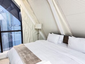 Una habitación de hotel con un ventanal grande, una cama con ropa blanca y una lámpara.