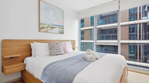 A hotel room with a large bed, a wooden headboard, a painting on the wall, and a view of a modern building through large windows. - The Roy Luxury 3BR with Huge Entertainer's Terrace (Melbourne)