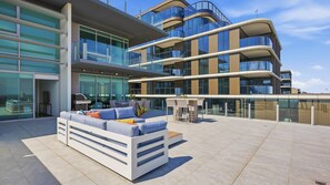 Ein modernes Gebäude mit Glasfassade, einer Dachterrasse mit Sofa und Stühlen, und wolkenlosem Himmel.
