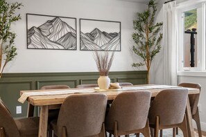 A dining area with a wooden table, six chairs, and two framed mountain landscape artworks on the wall.