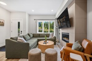 A modern living room with a sectional sofa, a round coffee table, and a leather armchair. A flat-screen TV is mounted on the wall, and a fireplace is visible. - Montane | Slope-Side | Hot Tub | Resort Access (Panorama)