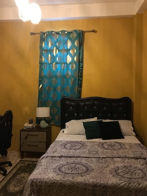 Ein Schlafzimmer mit Bett, Nachttisch, Lampe und Vorhang.