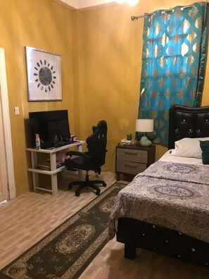 Ein Schlafzimmer mit Bett, Schreibtisch, Computer, Stuhl, Nachttisch und Vorhang.