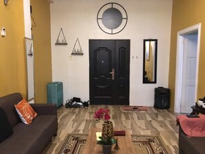 Een woonkamer met een bank, een bijzettafeltje met een plant, een klok aan de muur en een deur met een spiegel.