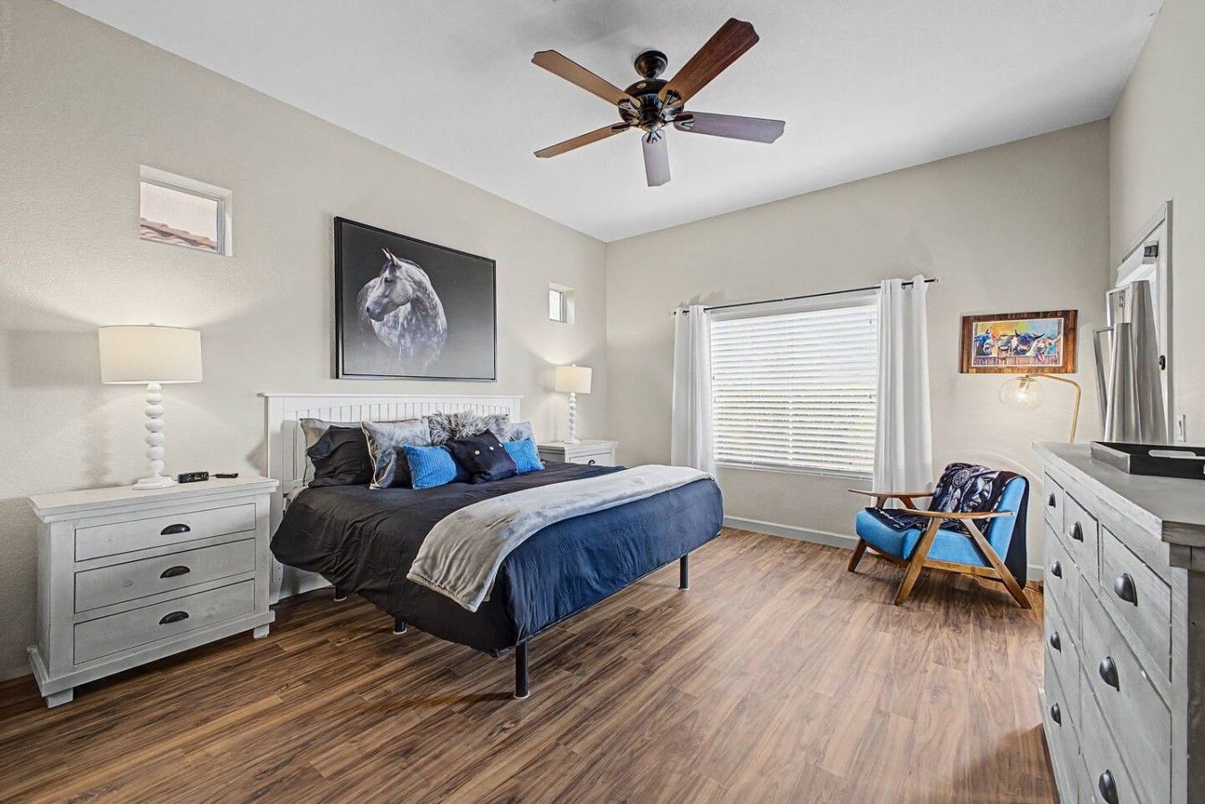 A bedroom with a bed, bedside tables, a ceiling fan, a window with blinds, and a chair.