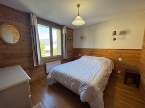 A bedroom with a wooden wall, a bed, a radiator, a window with curtains, and a ceiling light. - Duplex 8 pers - Vue montagne - Balcon sud - Pistes à 100 m (Vars)