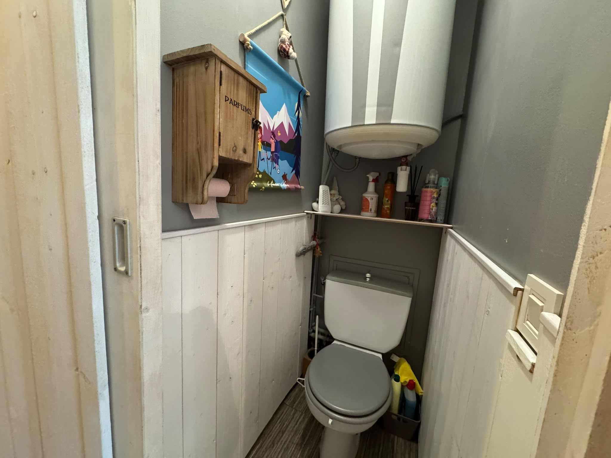 A compact bathroom with a toilet, a wooden cabinet labeled 'PARFUMS,' and a shelf with toiletries.
