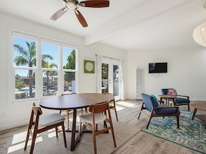 A spacious living room with a dining area, large windows, a ceiling fan, and a flat-screen TV.