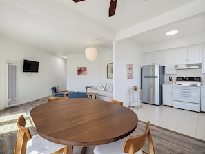 Uma cozinha moderna com eletrodomésticos de aço inoxidável, uma sala de jantar com uma mesa de madeira redonda e uma área de estar com sofá e televisão.