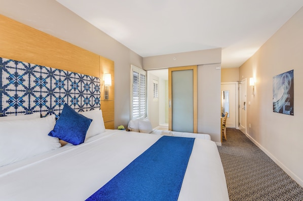 Een hotelkamer met een groot bed, een bekledingspaneel met een patroon, een blauwe plaid en een gang naar een badkamer.