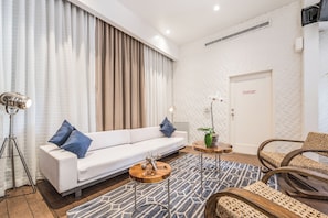 Een moderne woonkamer met een witte bank, een salontafel en een lamp. Er is een deur met een bordje met daarop een brandmeldpaneel.
