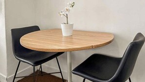Una mesa redonda de madera con dos sillas negras, un florero blanco con flores y una pared blanca lisa.