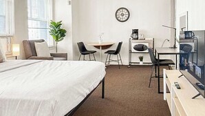 Una habitación de hotel con cama, escritorio, dos sillas, máquina de café, televisión y un reloj en la pared.