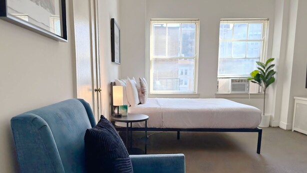A bedroom with a bed, a blue armchair, a bedside table with a lamp, and a window with blinds. - Gold Coast Hideaway | Vintage & Inviting Studio (Chicago)