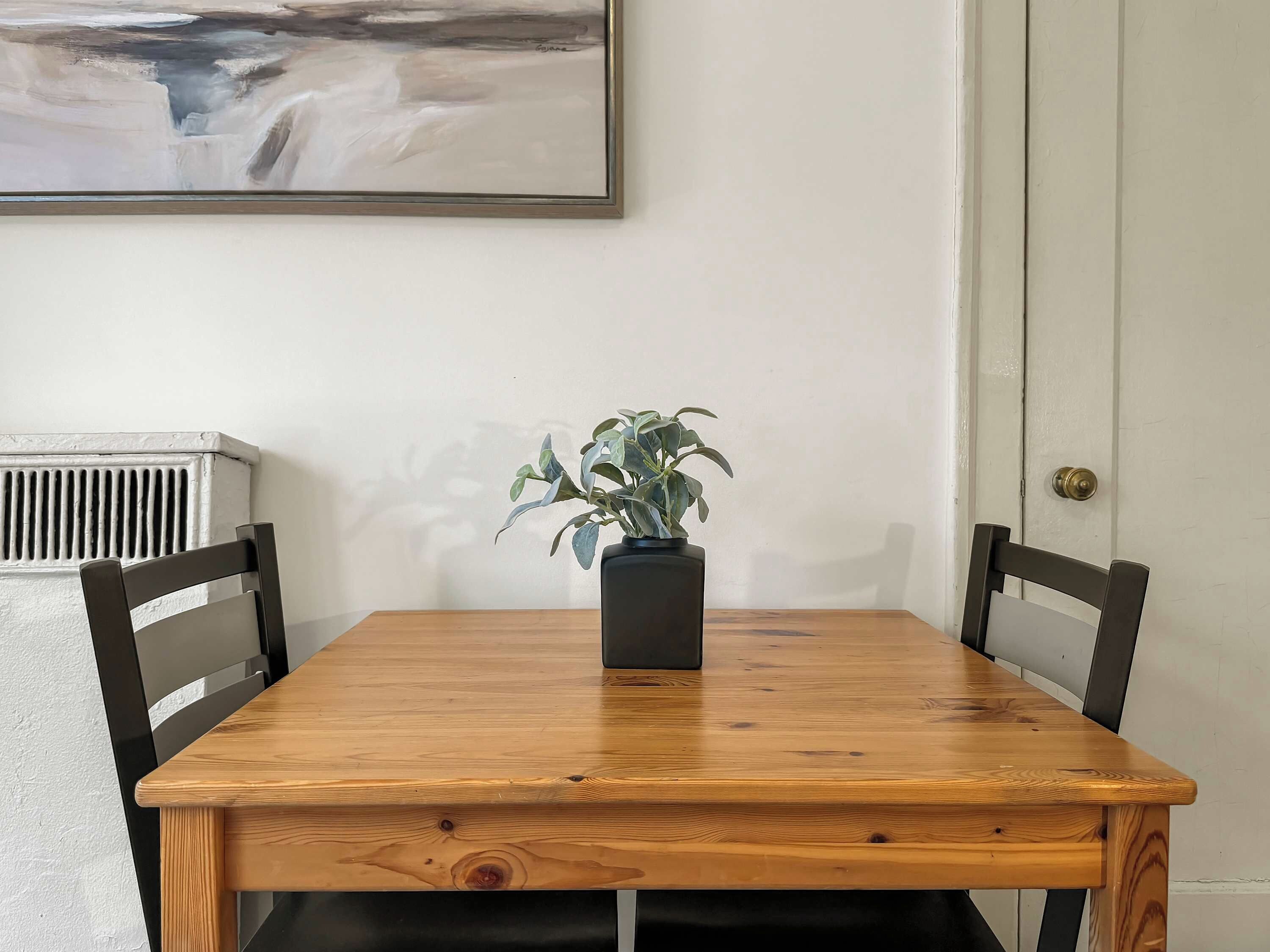Een houten eettafel met twee stoelen, een kamerplant en een schilderij aan de muur.