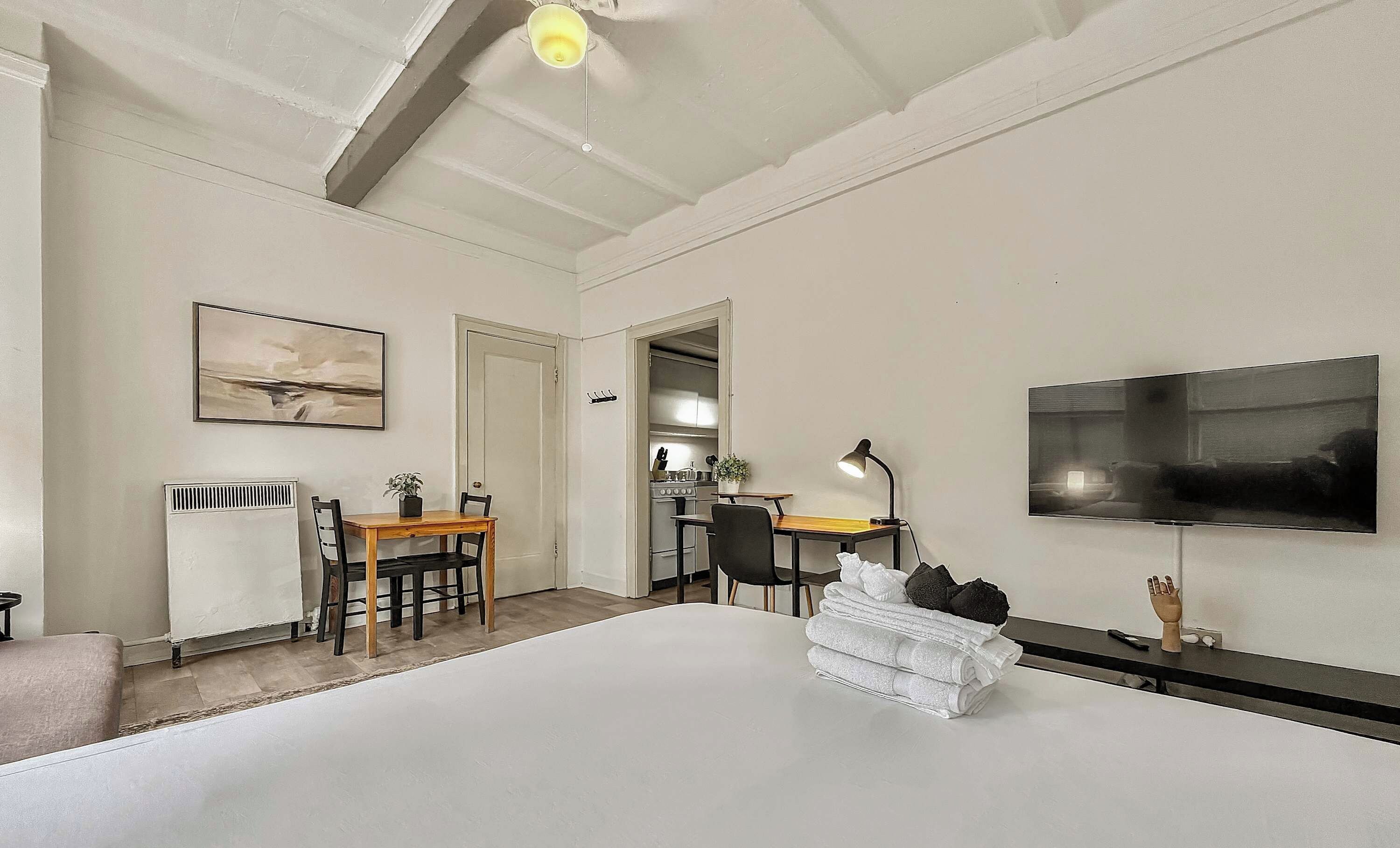 Een moderne hotelkamer met een groot bed, een bureau met stoel, een aan de muur bevestigde televisie en een keukenruimte zichtbaar door een open deur.