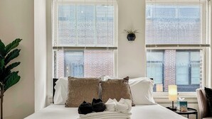 A neatly made bed with white and brown pillows, a stack of white towels, and a small table with a lamp and tissues. - Chic Studio in the Heart of the Gold Coast (Chicago)