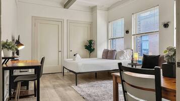 A bedroom with a bed, bedside tables, a desk, and a chair. There are windows with blinds, a ceiling fan, and a rug on the floor.