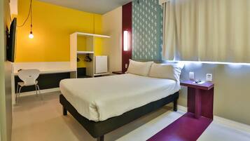 Quarto de hotel com cama, escrivaninha, cadeira, guarda-roupa e janela com cortinas.