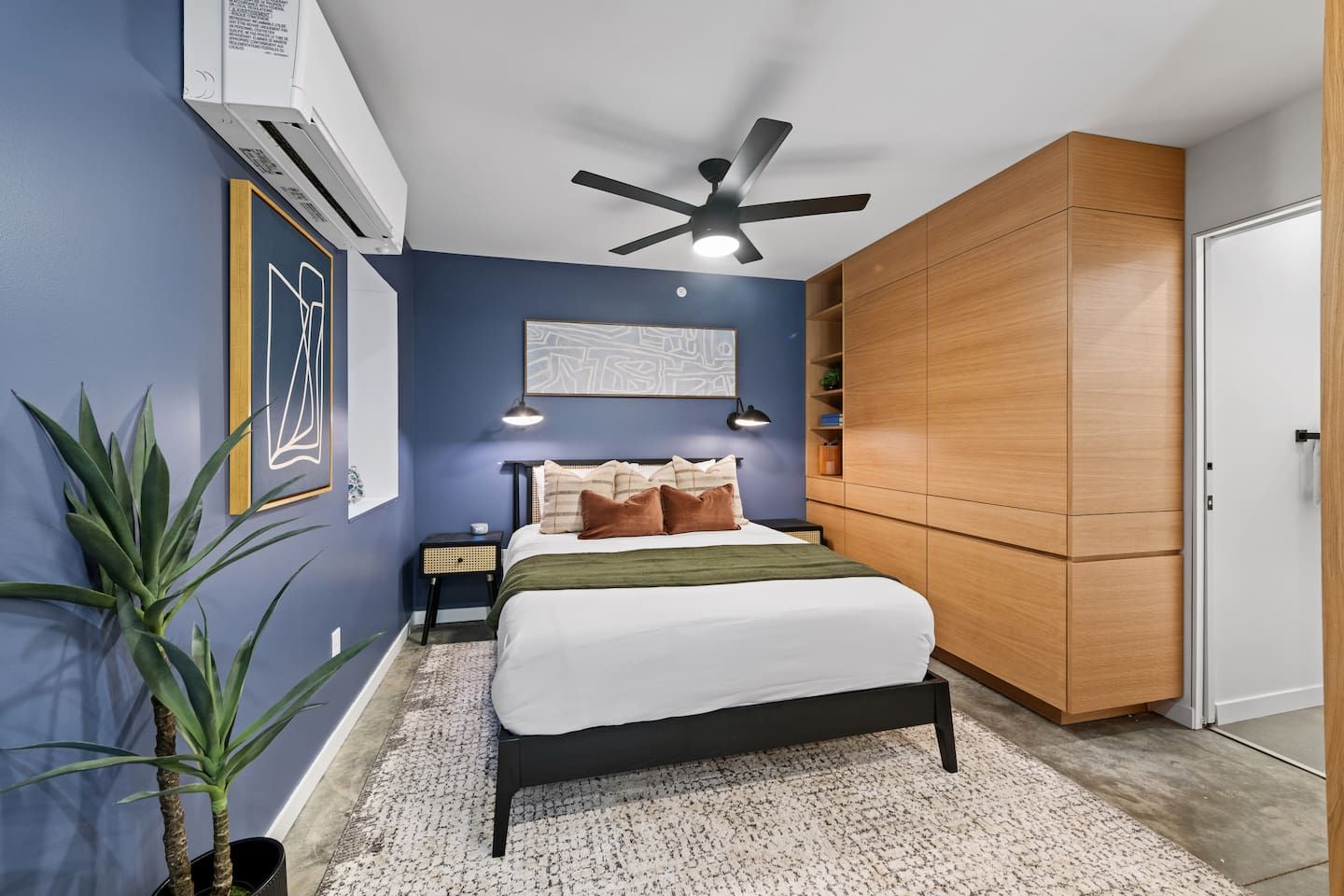 A bedroom with a bed, bedside tables, a ceiling fan, a wall-mounted air conditioner, and a large wardrobe.