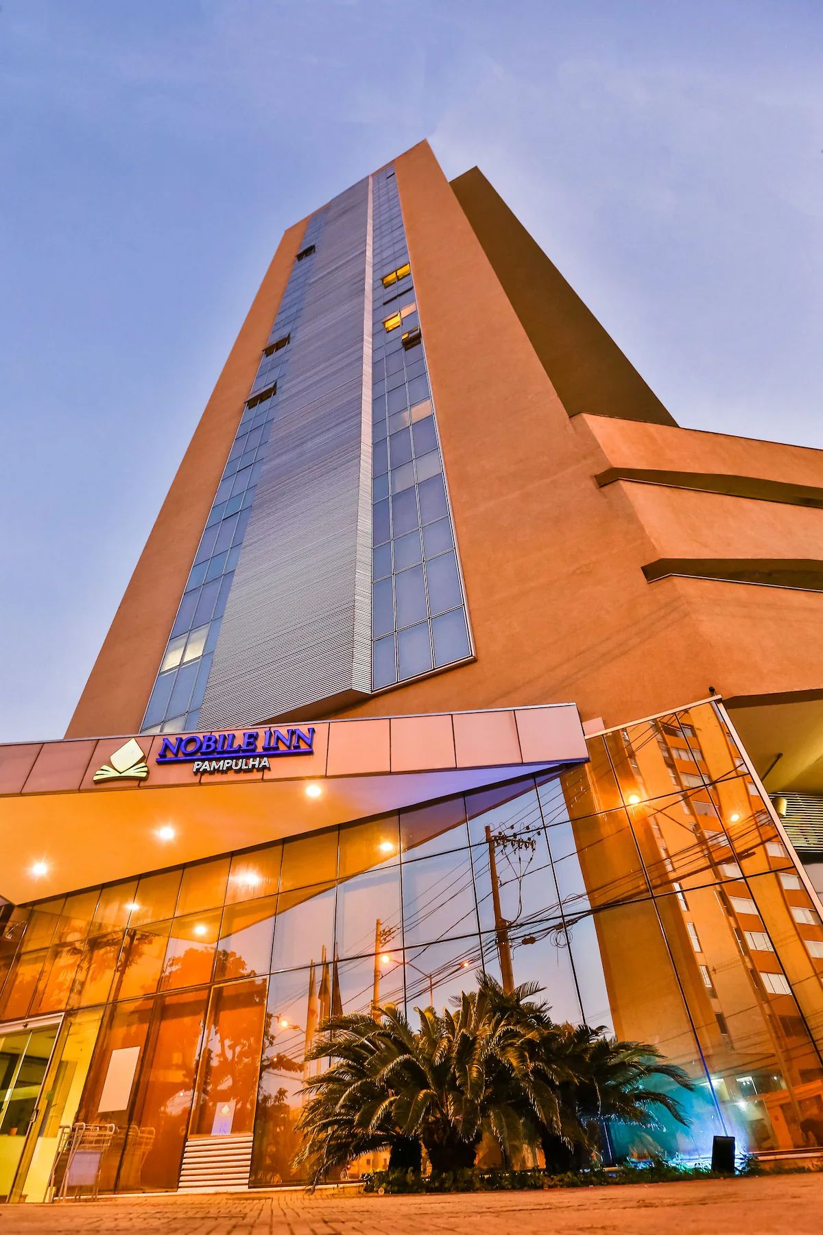Um edifício alto com design moderno, apresentando uma entrada destacada com portas de vidro e uma placa com a inscrição "NOBLE INN PAMPULHA".