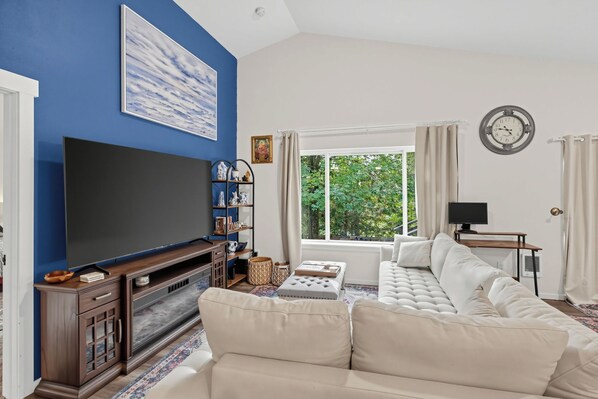A living room with a large flat-screen TV, a beige sectional sofa, a wooden entertainment unit, and a window with a view of greenery.