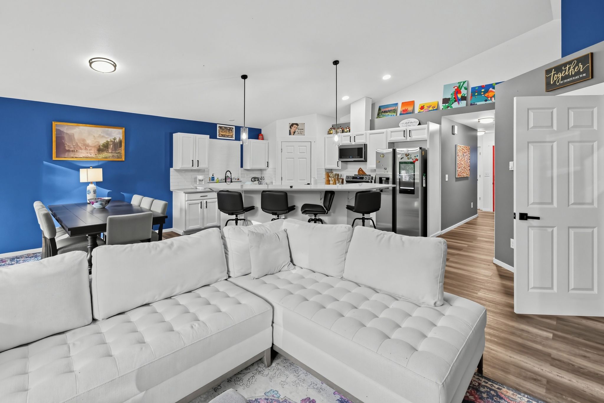 Una sala moderna con un sofá modular, un comedor con mesa y sillas, una cocina con armarios y electrodomésticos blancos, y un pasillo que conduce a otra habitación.