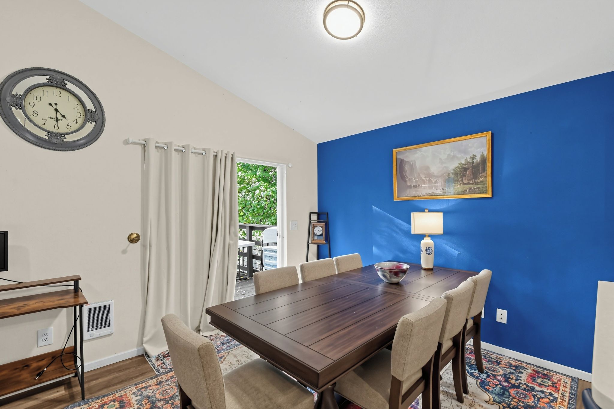 A dining area with a wooden table and chairs, a clock, a painting, and a lamp.