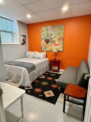 Ein Zimmer mit einem Bett, einem Schlafsofa, einem kleinen Tisch, einer Lampe und einem farbenfrohen abstrakten Gemälde an der Wand.