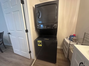 Una unidad compacta de lavadora y secadora con panel de control, ubicada en una esquina de una habitación donde se ve una puerta y una silla.