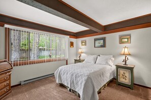 A bedroom with a large bed, two bedside tables with lamps, a window with blinds, and a view of greenery outside.