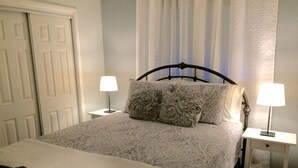 Una cama bien hecha con una colcha estampada y dos almohadas decorativas, flanqueada por dos mesitas de noche con lámparas, en una habitación con una puerta y una ventana con cortinas.