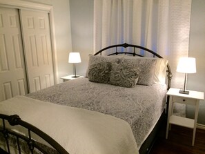 Un llit ben arrenjat amb un estovat estampat i dos coixins decoratius, flanquejat per dues taules de nit amb làmpades, en una habitació amb una porta i una finestra amb cortines.