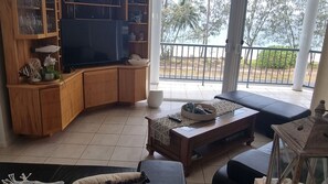 A living room with a television, wooden cabinets, a sofa, and a balcony with a view of trees. - Salt on South- Beach front views (South Mission Beach)