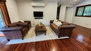 A living room with a brown leather sofa set, a coffee table with fruit, a flat-screen TV, and an air conditioning unit on the wall.