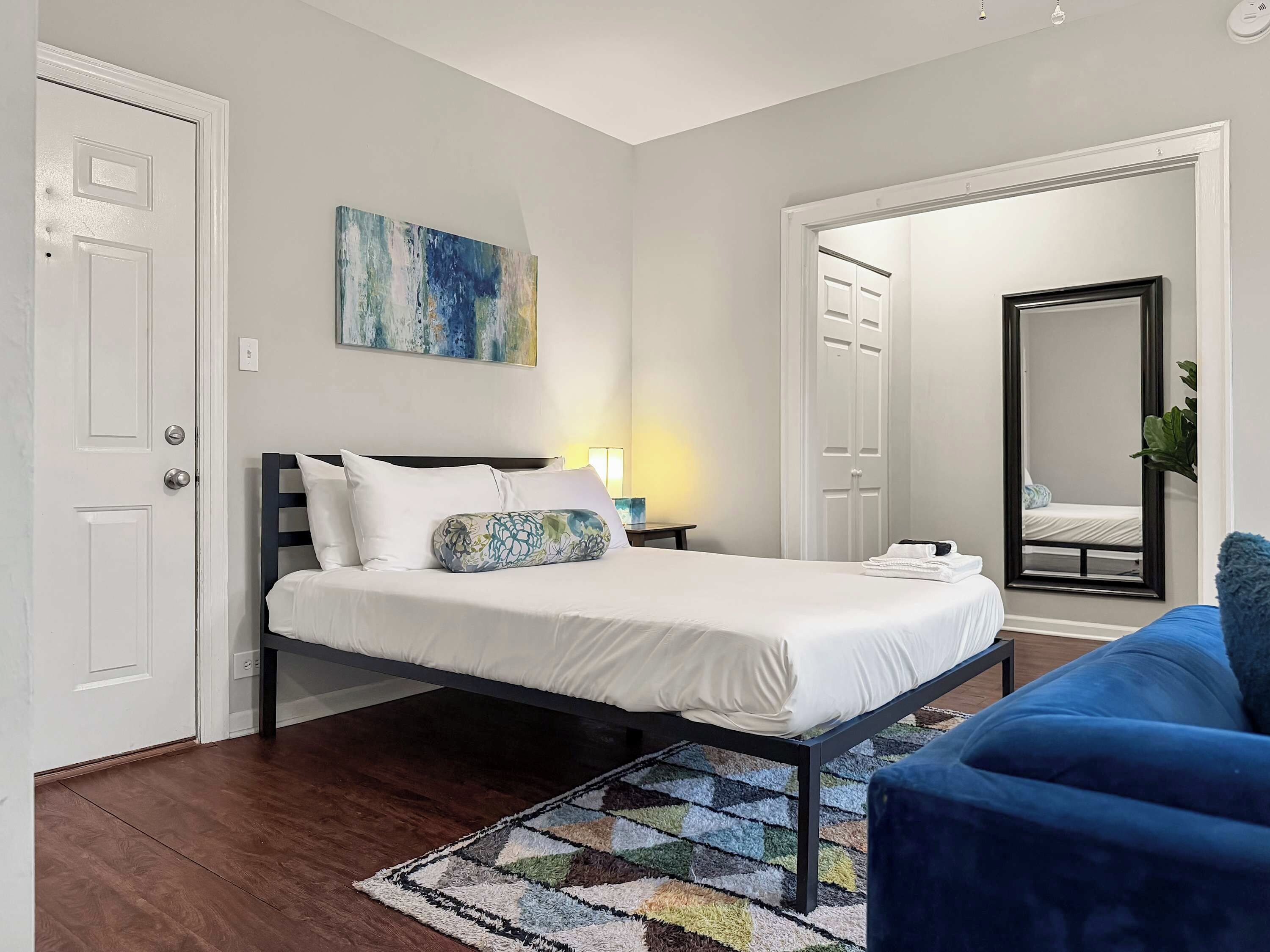 Un dormitorio ordenado con una cama, un sofá azul, un tapete estampado y un cuadro en la pared.