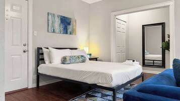 Un dormitorio ordenado con una cama, un sofá azul, un tapete estampado y un cuadro en la pared.