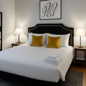 A neatly made bed with white linens and two mustard-colored pillows, flanked by two bedside lamps on wooden nightstands. - Boystown Chic 2BR Escape (Chicago)