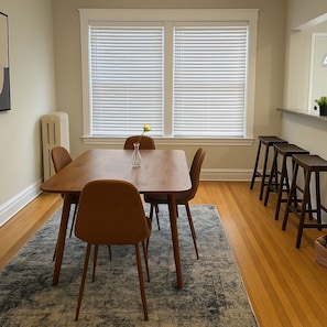 A dining area with a wooden table and chairs, a radiator, and a window with blinds. - Boystown Chic 2BR Escape (Chicago)