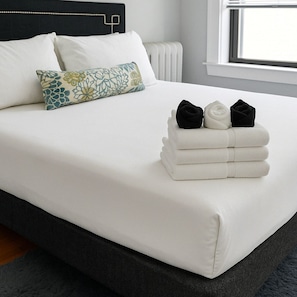 Een net opgemaakt bed met een witte deken, een decoratief kussen en een stapel opgevouwen handdoeken.