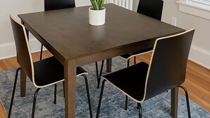 Uma sala de jantar com uma mesa retangular, quatro cadeiras, um vaso de planta, um radiador e uma janela com persianas.