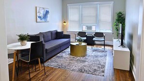 A modern living room with a grey sofa, a small round coffee table, and a white side table. There is a painting on the wall, a floor lamp, and a large window with blinds. - Chic & Fully Furnished 2BR Apartment (Chicago)