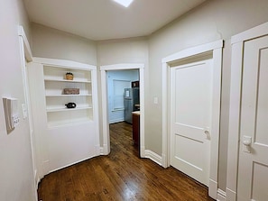 A hallway with white walls, wooden flooring, and white doors. Shelves with baskets and a clock are visible.