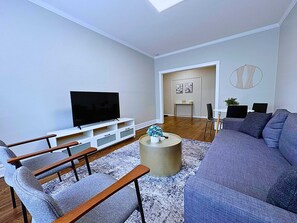 Una sala moderna con un televisor de pantalla plana sobre un soporte blanco, un sofá gris, una mesa de centro con objetos decorativos, y una zona de comedor con una mesa pequeña y sillas.