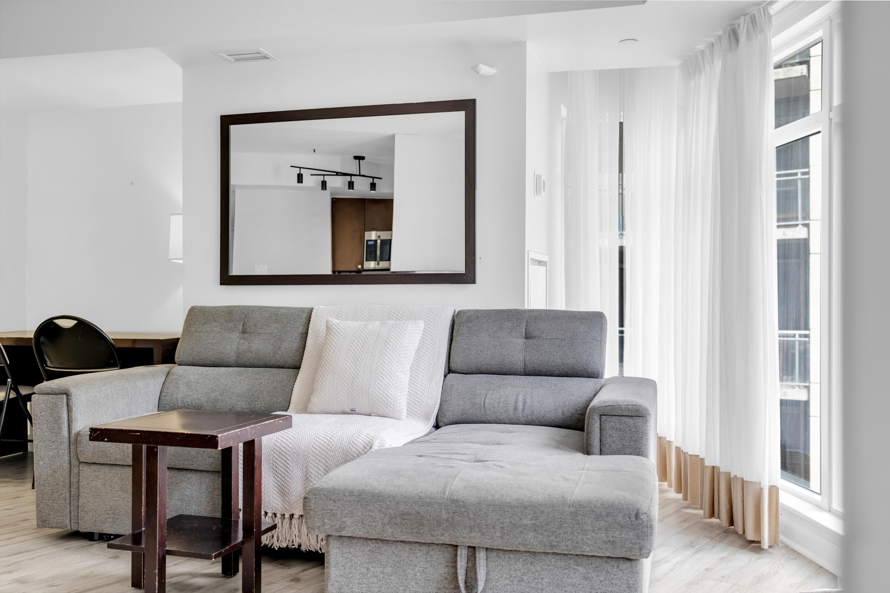 A modern living room with a grey sectional sofa, a small wooden coffee table, a large mirror, and a view of a kitchen through the mirror.