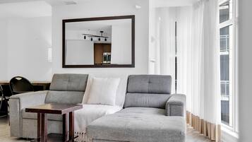A modern living room with a grey sectional sofa, a small wooden coffee table, a large mirror, and a view of a kitchen through the mirror.