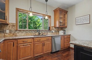 A kitchen with wooden cabinets, a stainless steel dishwasher, and a window with a view of greenery.