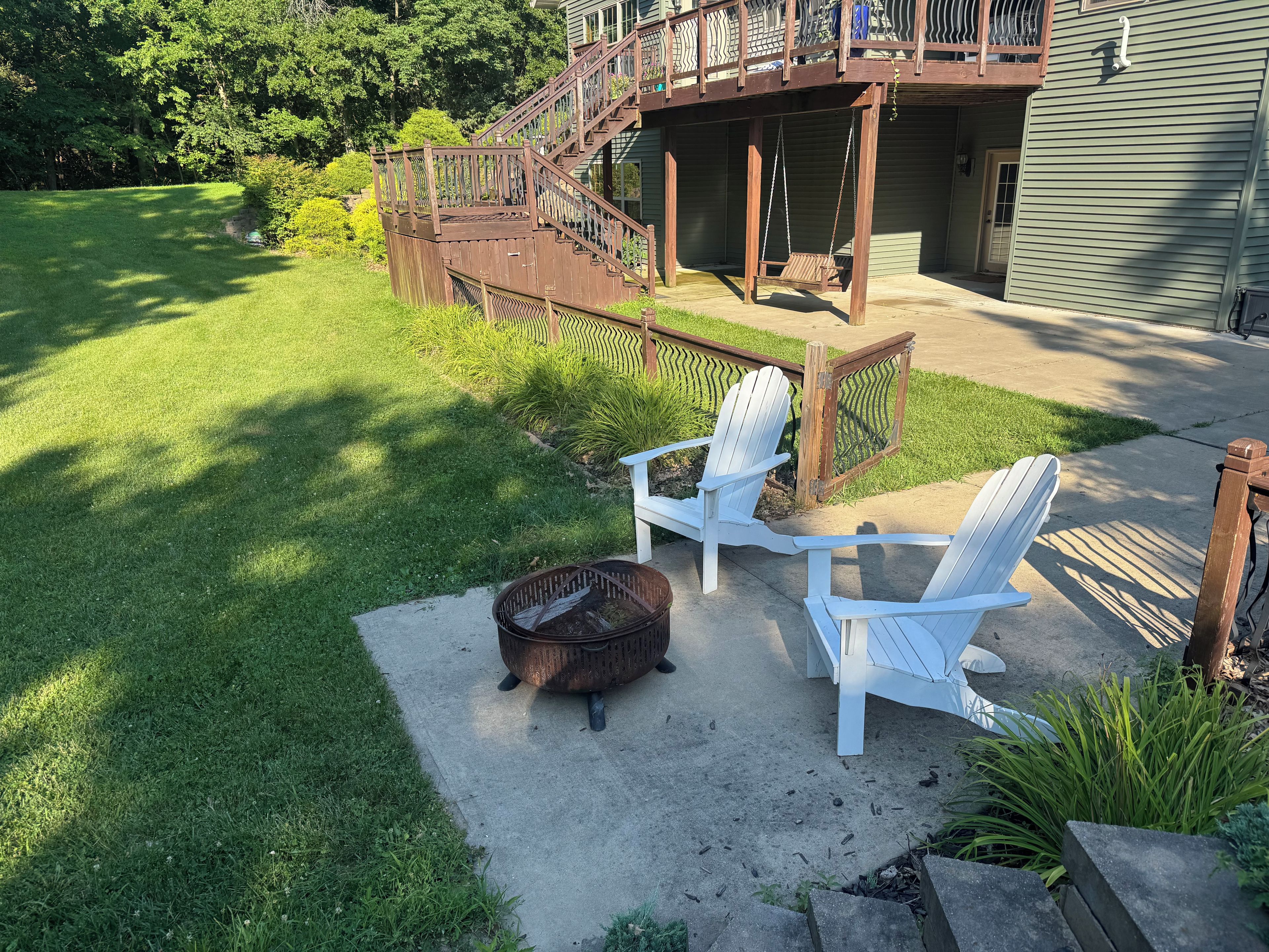 Ein Garten mit Feuerstelle, zwei Adirondack-Stühlen, einer Holzterrasse und einem Haus mit Balkon.